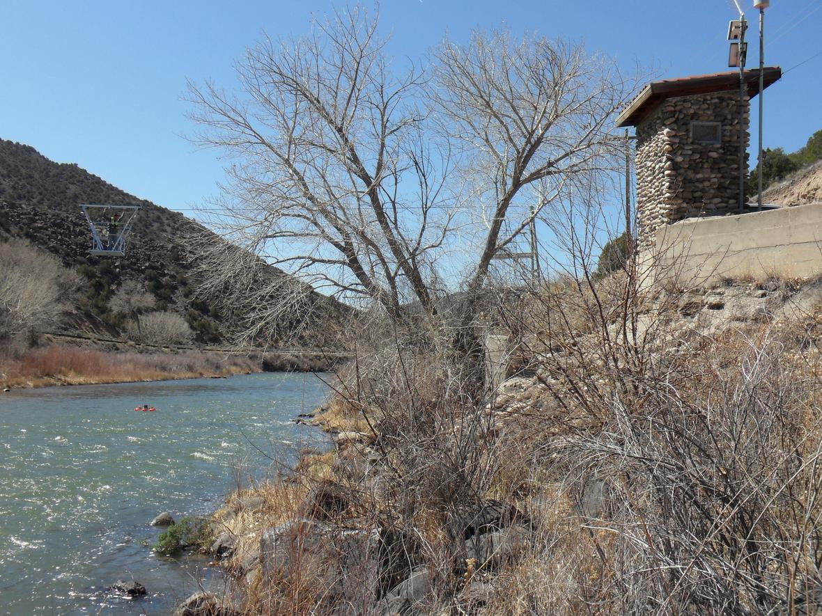 Image of streamgage on the Rio Grande at Embudo. Gage house made oflocal stone, cableway for measureing flow, riffles, and kayaker inriver.